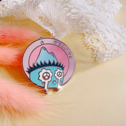 sad kappa (japanese water spirit) pin in front of white lace and pink foxtail plants. pin features crying turquoise kappa with big eyes on a pink circle with a light pink border. hiragana on the border says ‘sad face’.